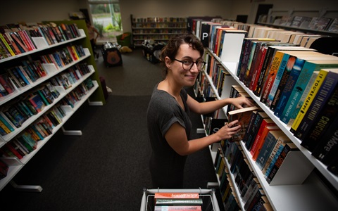 A young female places a book on a library shelf. 
